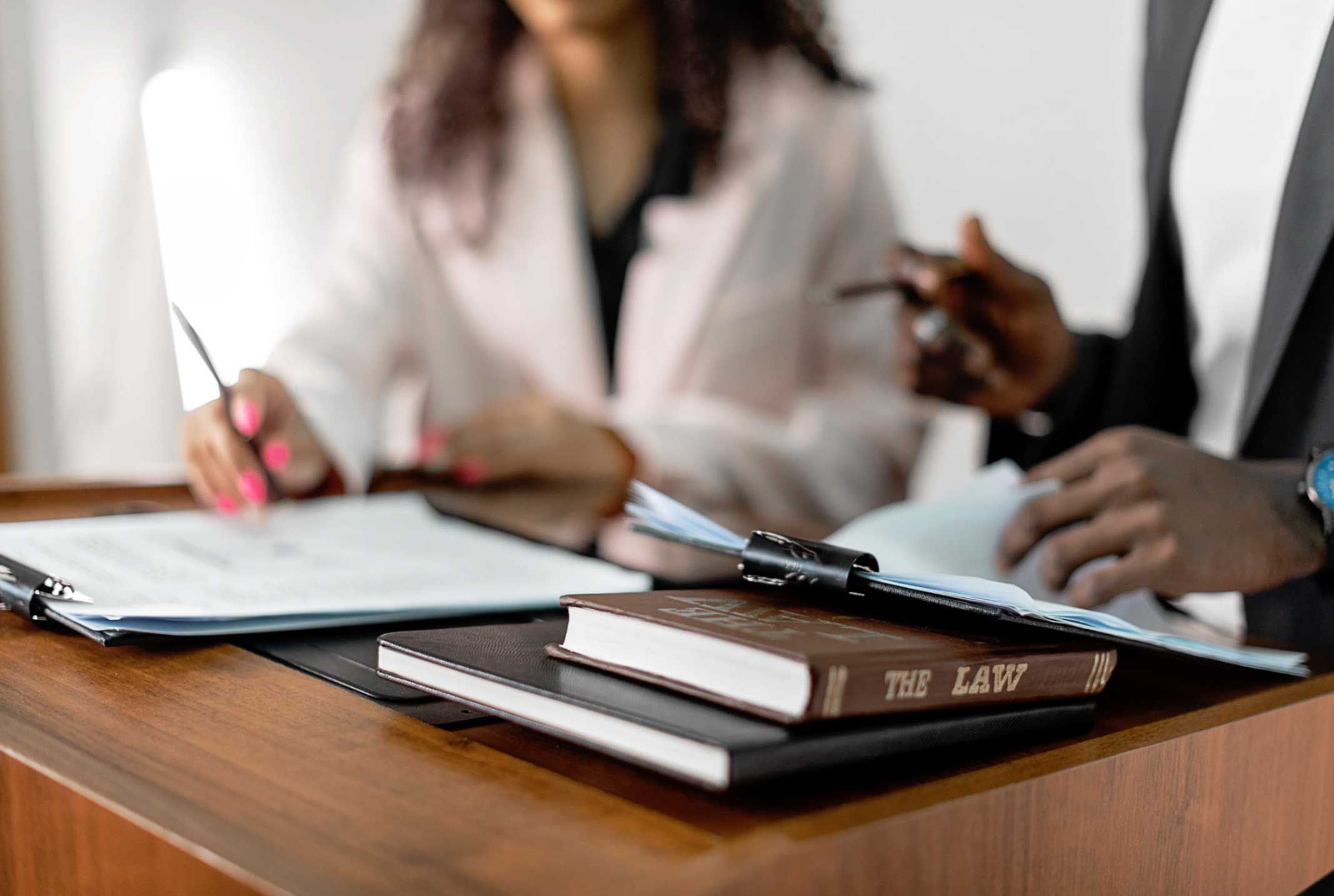 Two people review documents at a desk with a law book and a notepad in the foreground.