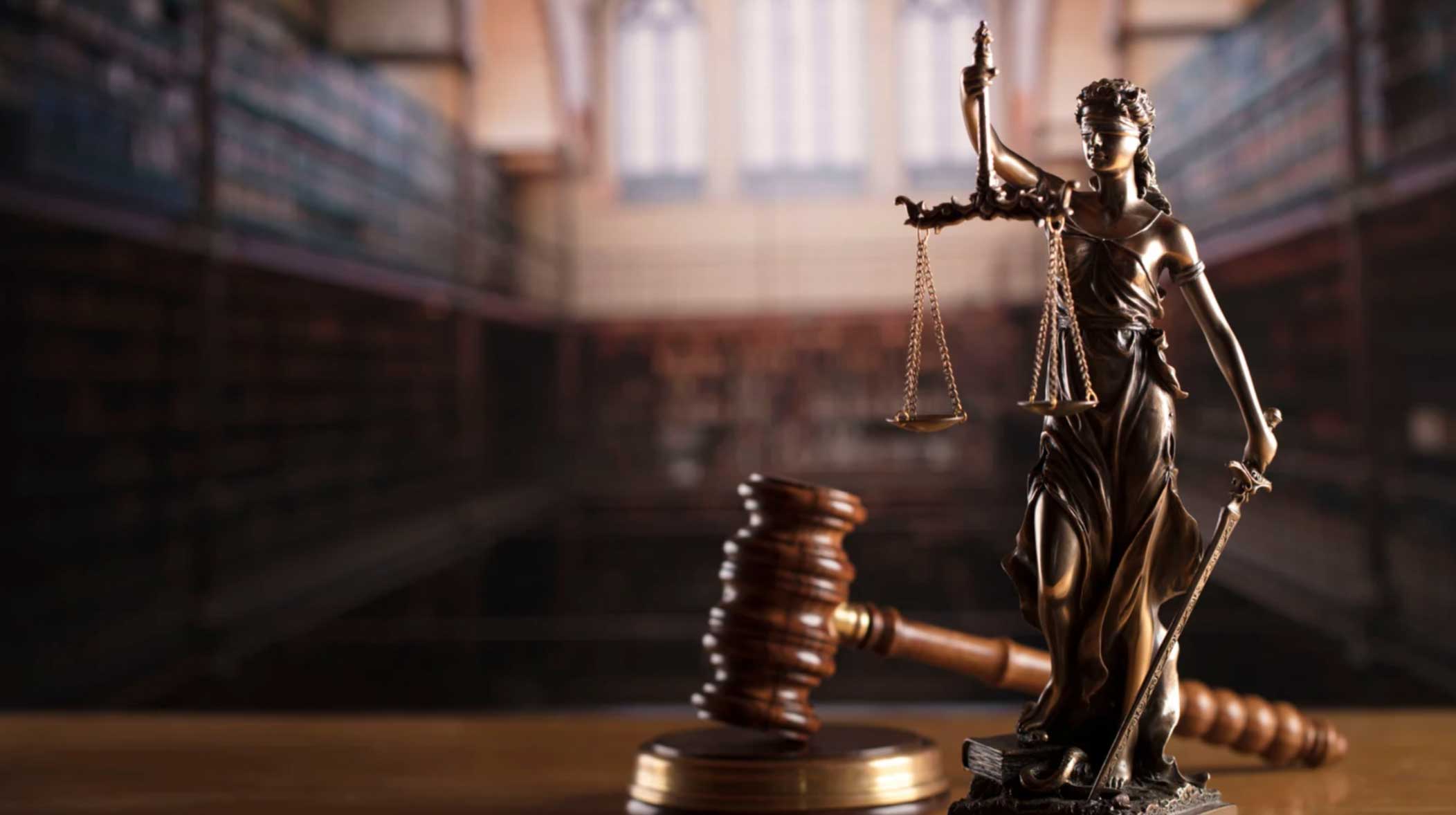 cta-bk-img Bronze Lady Justice statue and wooden gavel on a table in a blurred library or courtroom background.