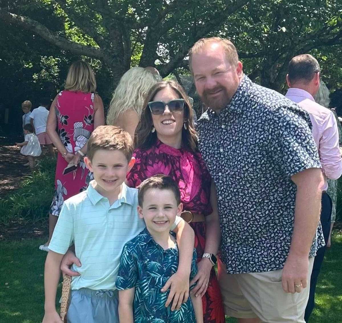 A family of four poses outside, smiling together on a sunny day with trees and people in the background.