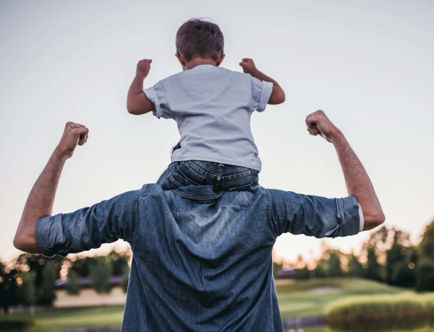 A man carries a child on his shoulders; both flex their arms outdoors at sunset.
