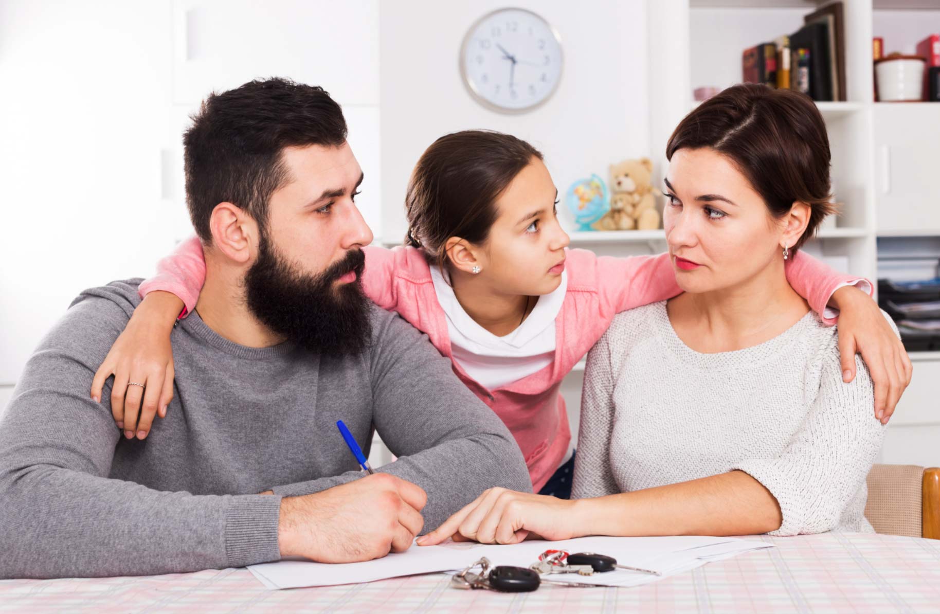 A man and woman talk seriously at a table while a girl hugs them, looking at the woman.