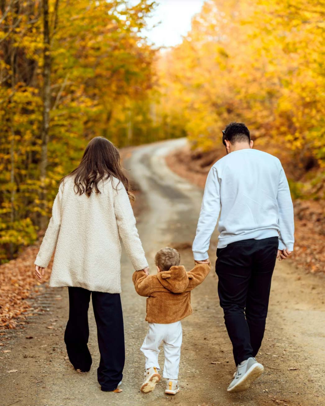 A family of three walks hand-in-hand down a forest path with autumn leaves.