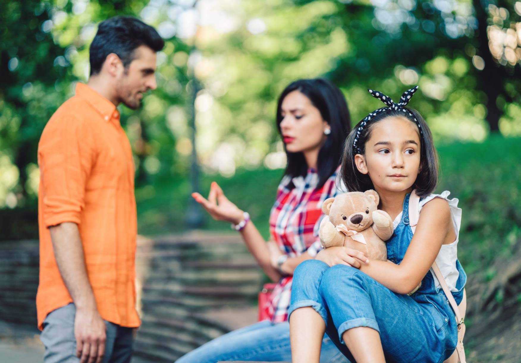 A sad girl holding a teddy bear sits apart as two adults argue in the background outdoors.