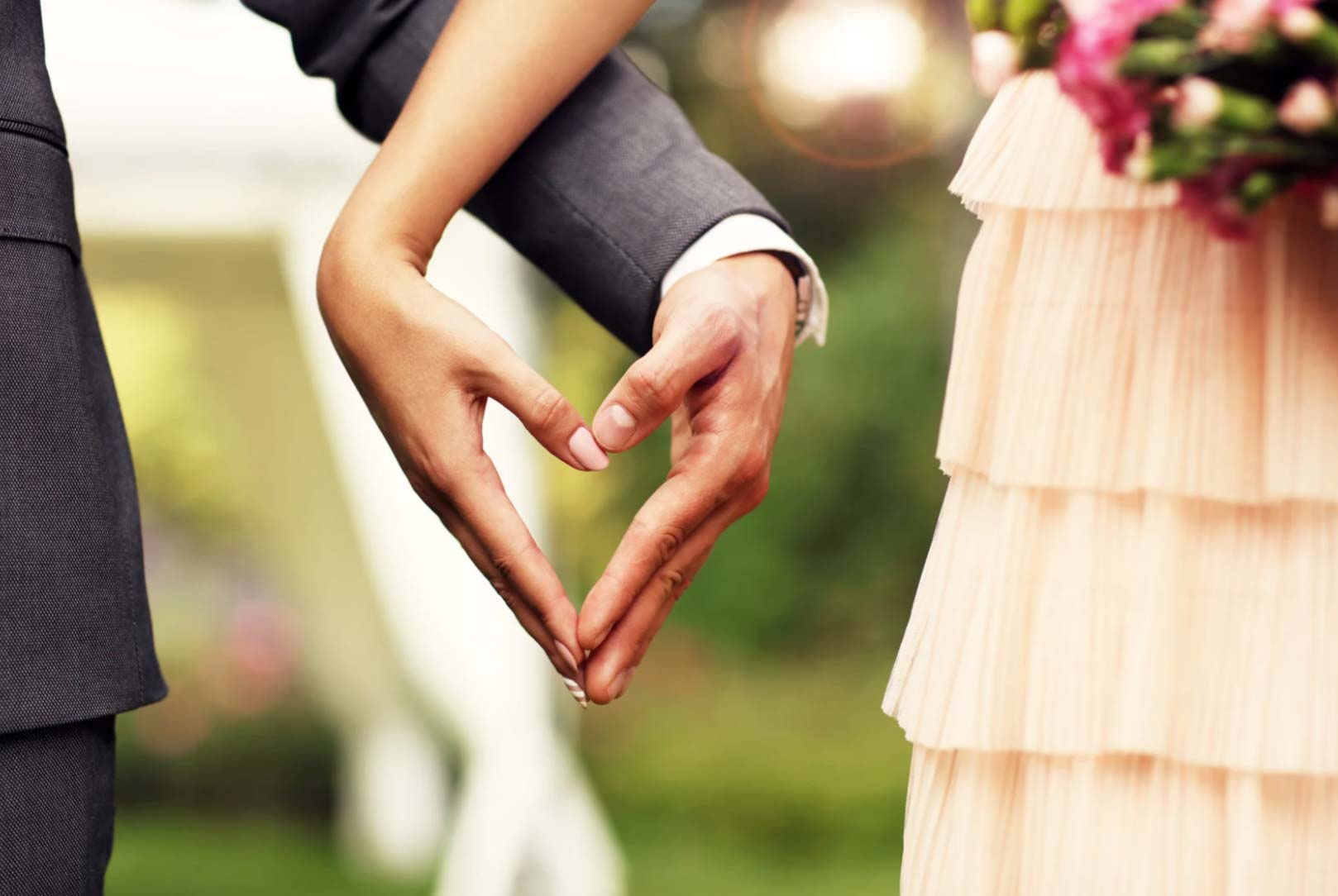 A couple forms a heart shape with their hands at an outdoor wedding, with a bouquet in view.