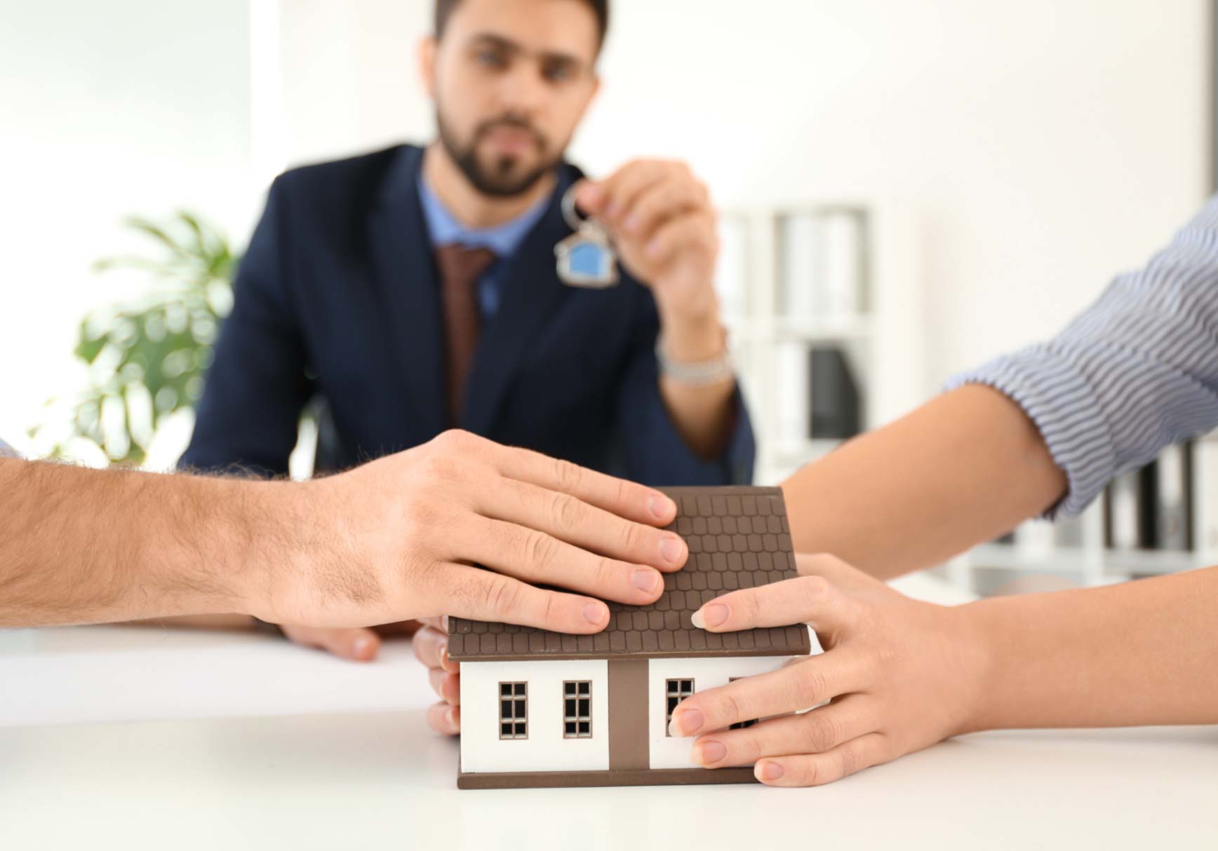 Three hands holding a model house, with a man in a suit holding keys in the background.